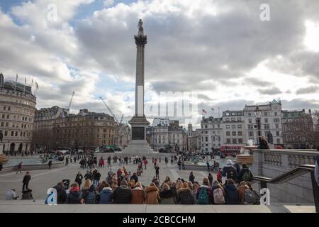 Group of young people seen sitting on the steps of Trafalgar Square London, with a general view towards Nelson's Column and surrounding buildings. Stock Photo