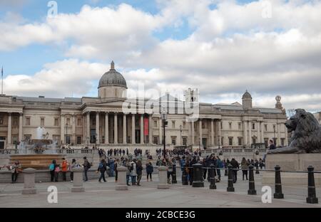 Tourists seen walking on the wide space of Trafalgar Square with the National Gallery museum in the background. Stock Photo