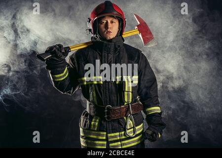 young caucasian handsome and strong fireman in uniform wearing helmet on head hold hammer in hands, going to save people from fire Stock Photo