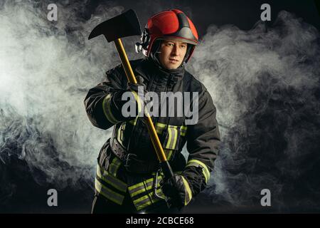 portrait of young caucasian fireman with hammer, wearing helmet and uniform isolated in black smoky background Stock Photo