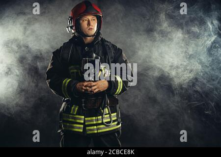 young fireman in protective coat using special equipment for fire fighting, isolated over smoky background Stock Photo