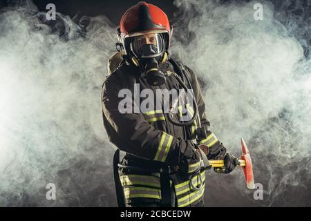 portrait of serious and confident caucasian fireman stand holding hammer, wearing special protective uniform in the smoky background Stock Photo