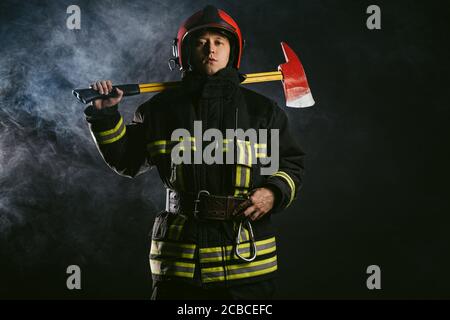 portrait of professional and confident fireman holding hammer, wearing protective uniform and helmet on head, stand isolated in smoky background Stock Photo