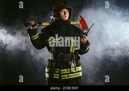 caucasian fire fighter using radio set at work, reporting about emergency situation standing isolated over smoky background Stock Photo