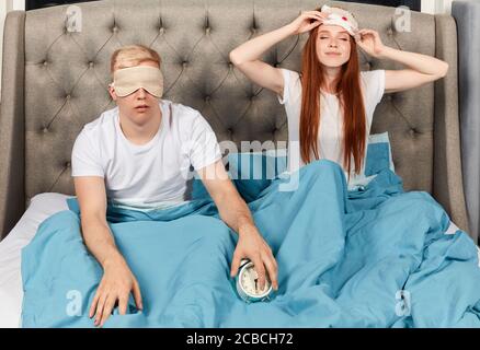young ginger woman is happy as she has a good night sleep, she is taking off sleeping mask, while her boyfriend sitting in mask and has problems with Stock Photo