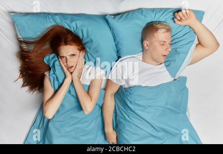 Unhappy ginger woman closing her ears with palms and her snoring husband lying in bedroom.close up top view photo Stock Photo