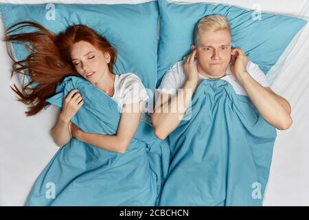 ginger girl snoring and angry man cannot sleep. nervous young blonde man closing his ears with fingers. he doesn't want to listen to his girl's snore. Stock Photo