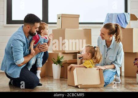 Caucasian family, man, woman and two girls play, smile with moving box. Father keep in hands youngest daughter, mother sat eldest daughter into box an Stock Photo