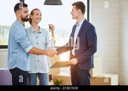 Young caucasian family and realtor conclude deal buy house. Men shake hands. Woman holds keys. Everyone smile. The sun shine. Stock Photo