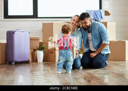 Kid wear denim overalls give little moving box parents who sit on floor and smile. Background moving boxes, suit and flower in pot Stock Photo