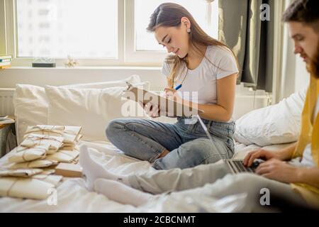 young caucasian married couple sort out letters invitations for housewarming. beautiful woman and handsome man sit with laptop at home, a lot of lette Stock Photo