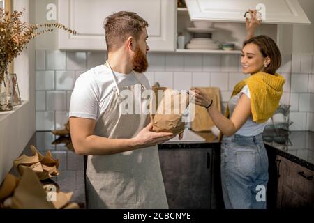 young married couple unpacking boxes after moving into a new apartment, they hold tableware in hands, woman arranges it in their new kitchen Stock Photo