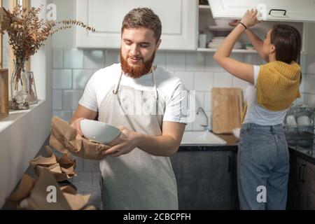 moving day of happy young married couple unpacking cardboard boxes, standing together in kitchen, unpacking carton package, holding tableware in hands Stock Photo