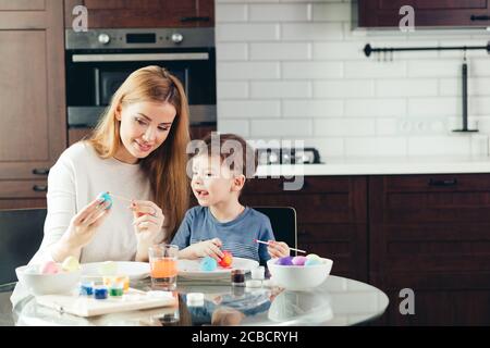 Concept of family holiday leisure in the kitchen. Beautiful Mom teaching her little boy child to color the Easter eggs.