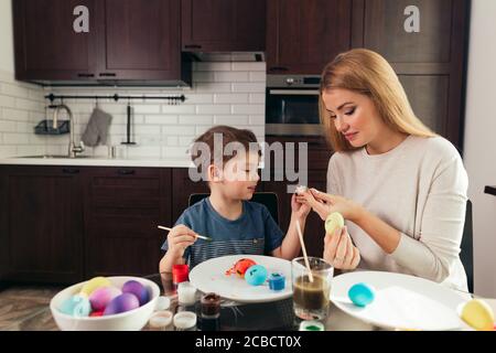 happy blonde mother and four-year old son making homemade Easter eggs, enjoying leisure time spent together at home. Family celebrating Easter. Parent Stock Photo