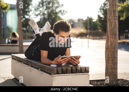 Young man watching movie on digital tablet while lying on bench at college campus Stock Photo