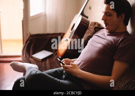 Young man yawning while using smart phone sitting at home Stock Photo