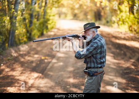 Side view on senior hunter man aims at trophy bird in autumn sunny forest, pointing gun at bird. Stock Photo