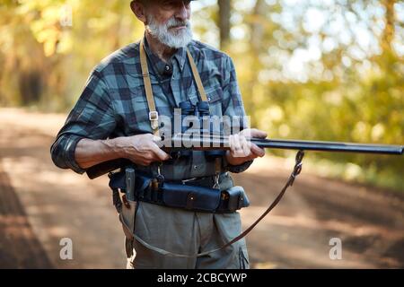 Older hunter man with grey beard holding gun to hunt on birds, ready to shoot. Forest background Stock Photo