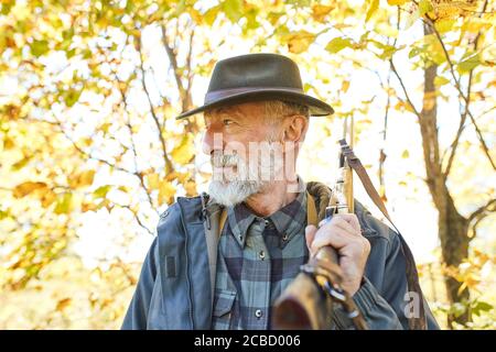Bearded senior hunter wearing hat stands with gun on shoulder, looks into the distance in search of trophy. Wild animals prey in hunting Stock Photo