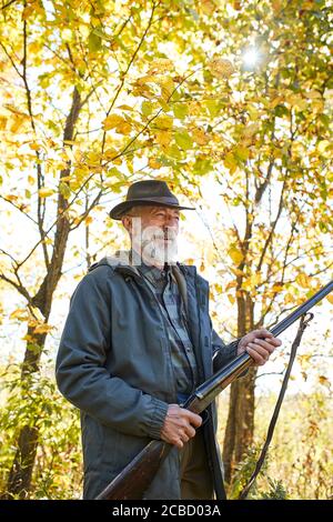 Smiling hunter male stand holding gun and look away, wearing hat and coat, autumn Stock Photo