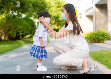 Asian mother help her daughter wearing medical mask for protection Covid-19 or coronavirus outbreak in village park to prepare go to school when back Stock Photo