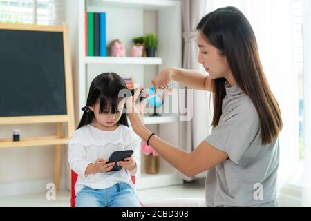 Asian Mother cutting hair to her daughter in living room at home while stay at home safe from Covid-19 Coronavirus during lockdown. Self-quarantine an Stock Photo