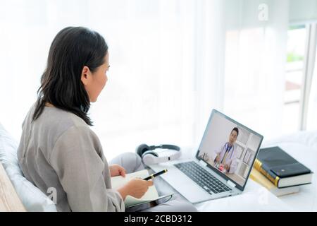 Back view of Asian woman making video call with her doctor with her feeling sick on laptop in bedroom for online healthcare digital technology service Stock Photo