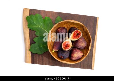 Fresh ripe figs, with one fig cut in half, in a wooden bowl on dark board with a green fig leaf. Healthy Mediterranean fig fruit on a white background Stock Photo