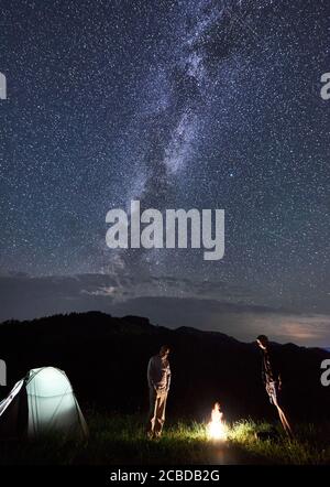 Couple of friends have a rest around a campfire near illuminated tourist tent in the mountains in summer, enjoying incredible weather under beautiful sky full of stars, Milky way. Astrophotography Stock Photo