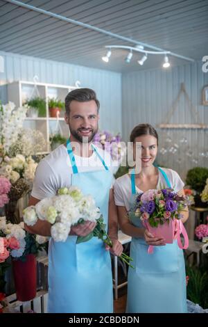 Bearded male and brown-haired female holding bouquet and box of flowers, smiling Stock Photo