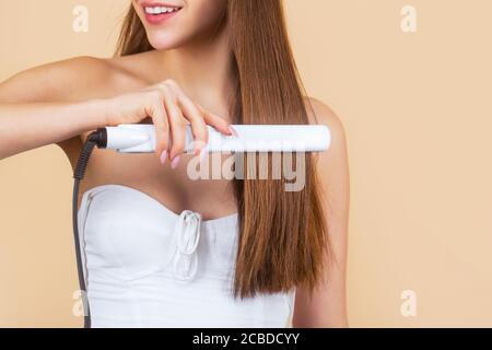 Smiling woman ironing long hair with flat iron. Happy young woman straightening hair with straightener. Portrait of young beautiful girl using styler Stock Photo