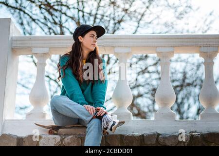 A beautiful hipster teenage girl sitting relaxed on a skateboard and looking into the distance. In the background is a White balustrade and a tree. Cl Stock Photo