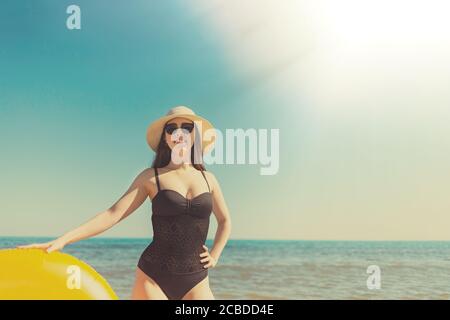 A young woman in a black swimsuit, sunglasses and hat holds an orange swimming circle with her hand and smiles. In the background sea and sky.The conc Stock Photo