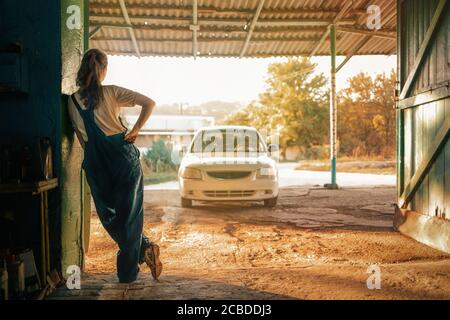 The concept of small business, feminism and women's equality. Young woman mechanic resting leaning against the wall. In the background, a car and a st Stock Photo