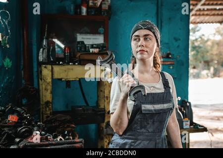 The concept of small business, feminism and women's equality. A young woman in overalls poses with a large wrench and looks away. Close up. Stock Photo