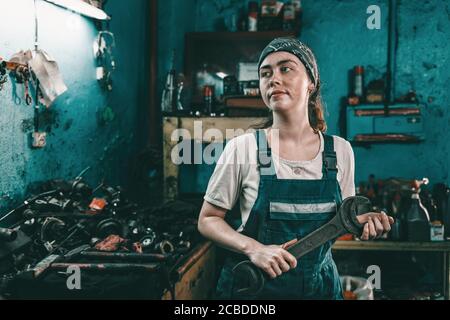 The concept of small business, feminism and women's equality. A young woman in overalls holds a large wrench and looks away. Stock Photo