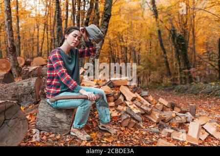 Preparation for the heating season. A young tired woman with an axe in her hands sits on a log and takes off her cap. There's a pile of firewood nearb Stock Photo