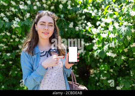 A beautiful young woman shows her index finger on a mobile phone. Green plants on a background. Copy space and mock up. Concept of modern technology a Stock Photo