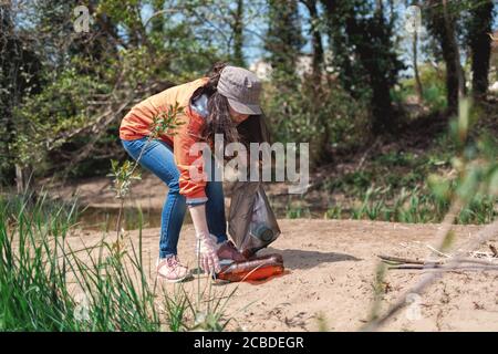 Woman volunteer picks up a plastic bottle on the sandy Bank of the river. Concept of Earth Day and environmental protection. Stock Photo