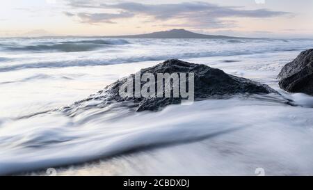 Waves crashing over rocks at sunrise with Rangitoto island in the distance. Image taken using slow shutter speed with the effect of silky water effect Stock Photo
