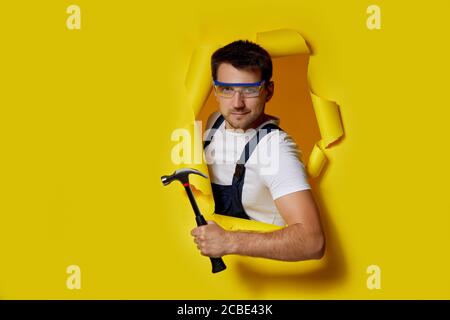 caucasian repairman worker in safety glasses and uniform holding hammer through hole in yellow paper. Stock Photo
