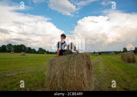 A handsome young man is sitting on a haystack against a blue cloudy sky. Stock Photo