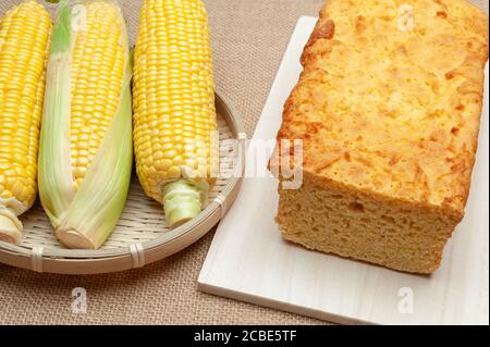 Homemade Brazilian corn bread on a cutting board and raw corn cobs in a woven bamboo basket. Isolated on jute background. Close-up. Stock Photo