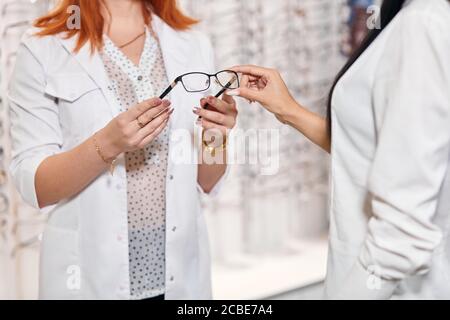young female doctor giving glasses to brunette woman, close up photo. client choosing a frame for eyeglasses