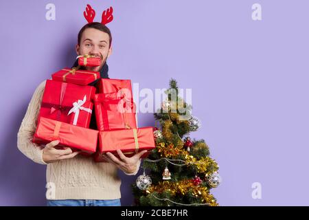Young man carries a lot of presents, various gift boxes in hands of handsome unshaven male, standing near small beautiful Christmas tree, isolated on Stock Photo