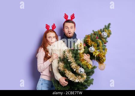 Pretty young couple holding small Christmas tree standing behind decorated for with worrying faces, looking at camera, scared by unexpected surprise, Stock Photo