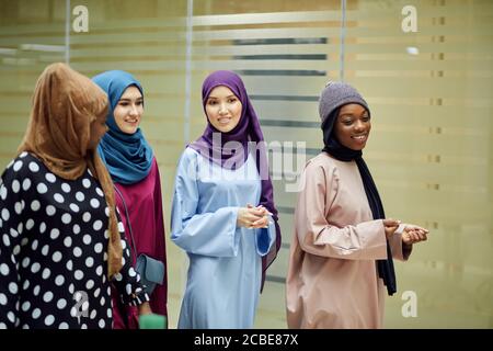 A group of four young muslim multiethnic business women dressed in fashionable tradition dresses chatting and walking together in business centre Stock Photo