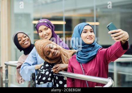 Cheerful muslim multiracial student girls wearing hijabs and long traditional wear having fun after classes and taking selfie at university hall. Stock Photo