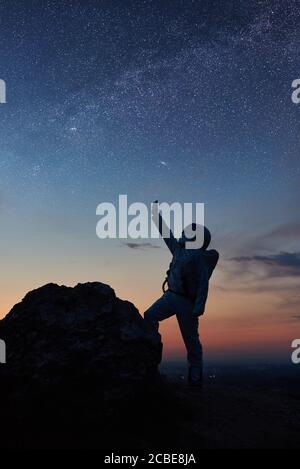 Space traveler standing on rocky mountain and pointing at stars. Silhouette astronaut wearing white space suit while looking at fantastic starry sky. Concept of galaxy, human in space and milky way. Stock Photo